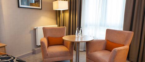 Habitación de hotel con dos sillones a cuadros, una mesa redonda con lámpara, cama con colcha estampada y ventana con cortinas.