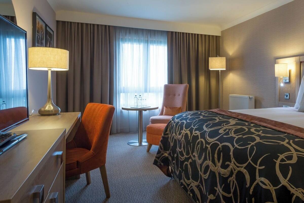 A hotel room with a bed, a desk, a chair, a table, and two lamps.
