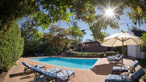 A swimming pool with lounge chairs and umbrellas, surrounded by greenery and a brick patio. - At Winkfield House (Cape Town)