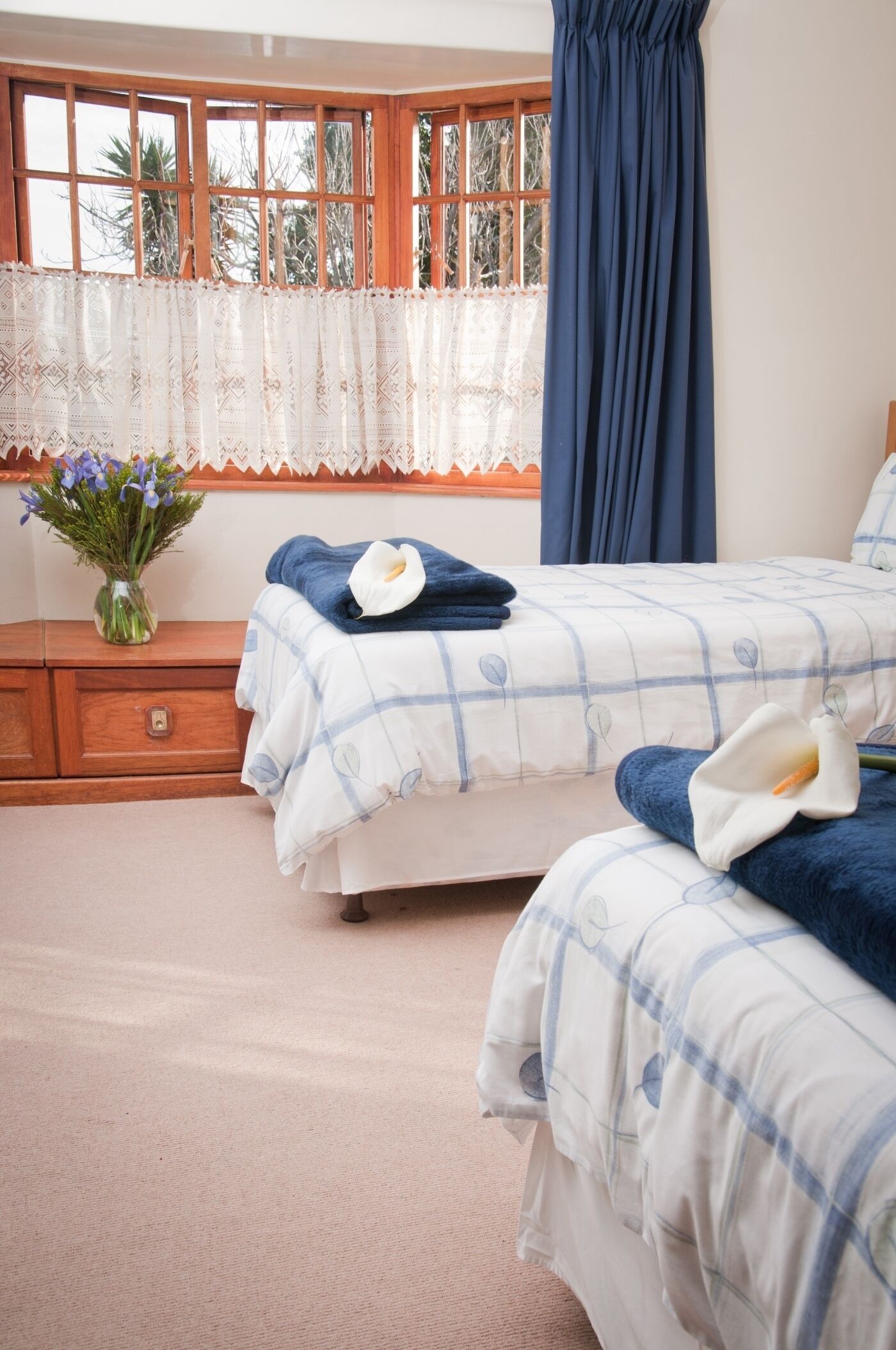 A bedroom with two beds, a wooden nightstand with a vase of flowers, and a window with lace curtains.