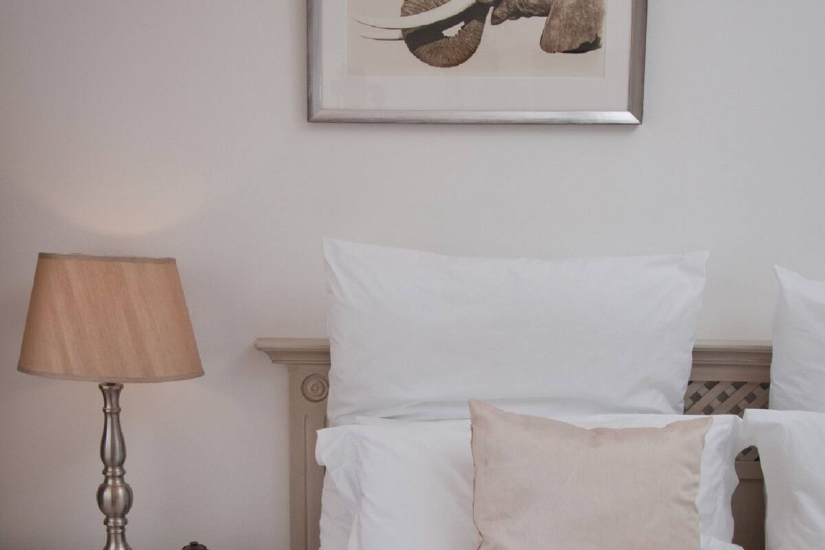 A neatly made bed with white linens and a patterned throw, a bedside lamp, a small decorative item, and a framed elephant artwork on the wall.