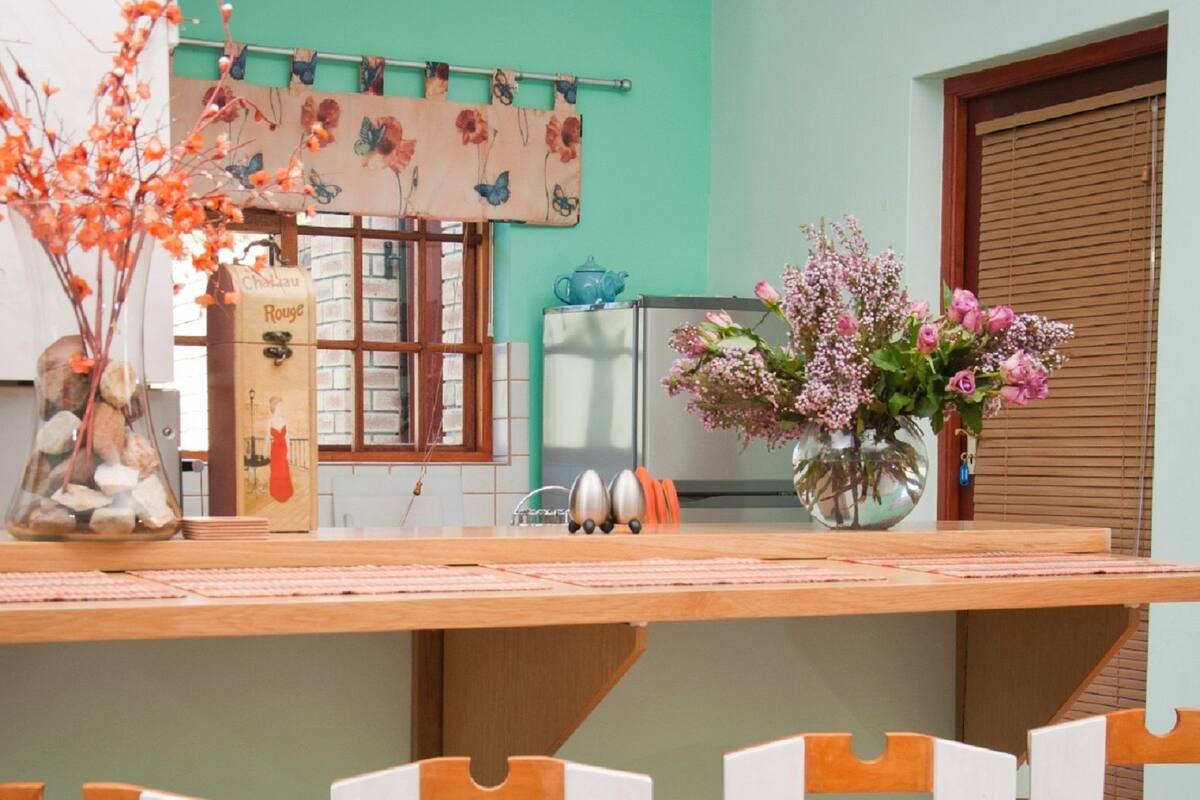 A kitchen with a wooden counter, white chairs, a clock on the wall, and a vase with flowers on the counter.
