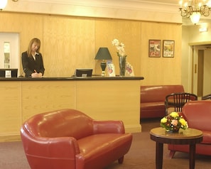 A hotel reception area with a receptionist, a reception desk, a green lamp, a vase with flowers, and a seating area with red leather chairs and a table. - Britannia Bournemouth (Bournemouth)
