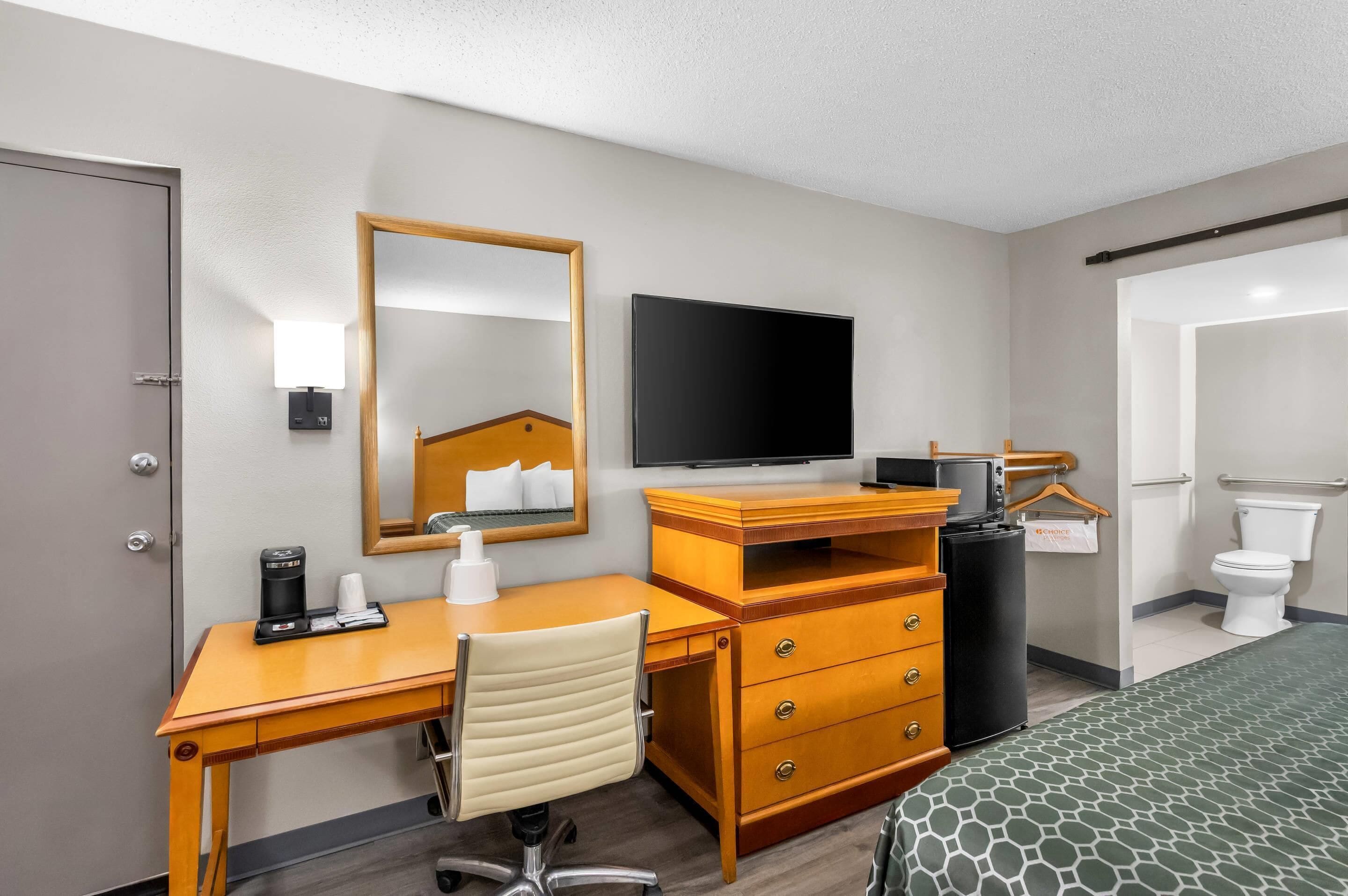 A hotel room with a wooden desk, a chair, a bed with a patterned bedspread, a television, a mirror, a bedside table with a lamp, and a trash can.