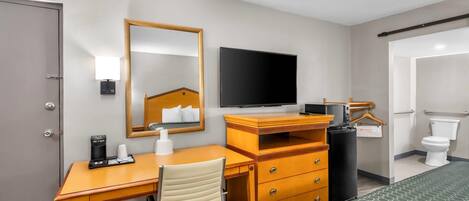 A hotel room with a wooden desk, a chair, a bed with a patterned bedspread, a television, a mirror, a bedside table with a lamp, and a trash can.