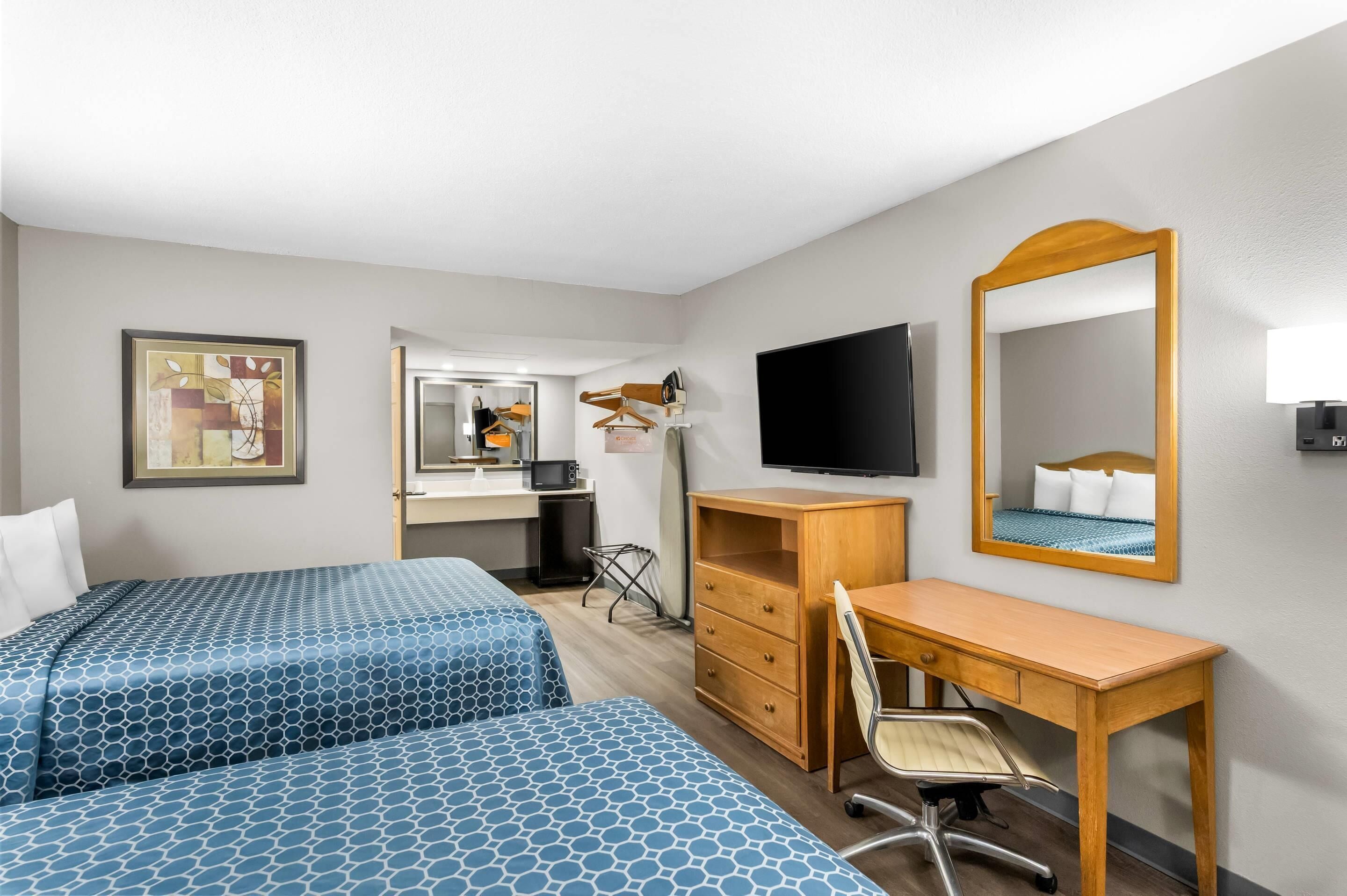 A hotel room with two beds, a desk, a chair, a TV, a mirror, and a small kitchenette.