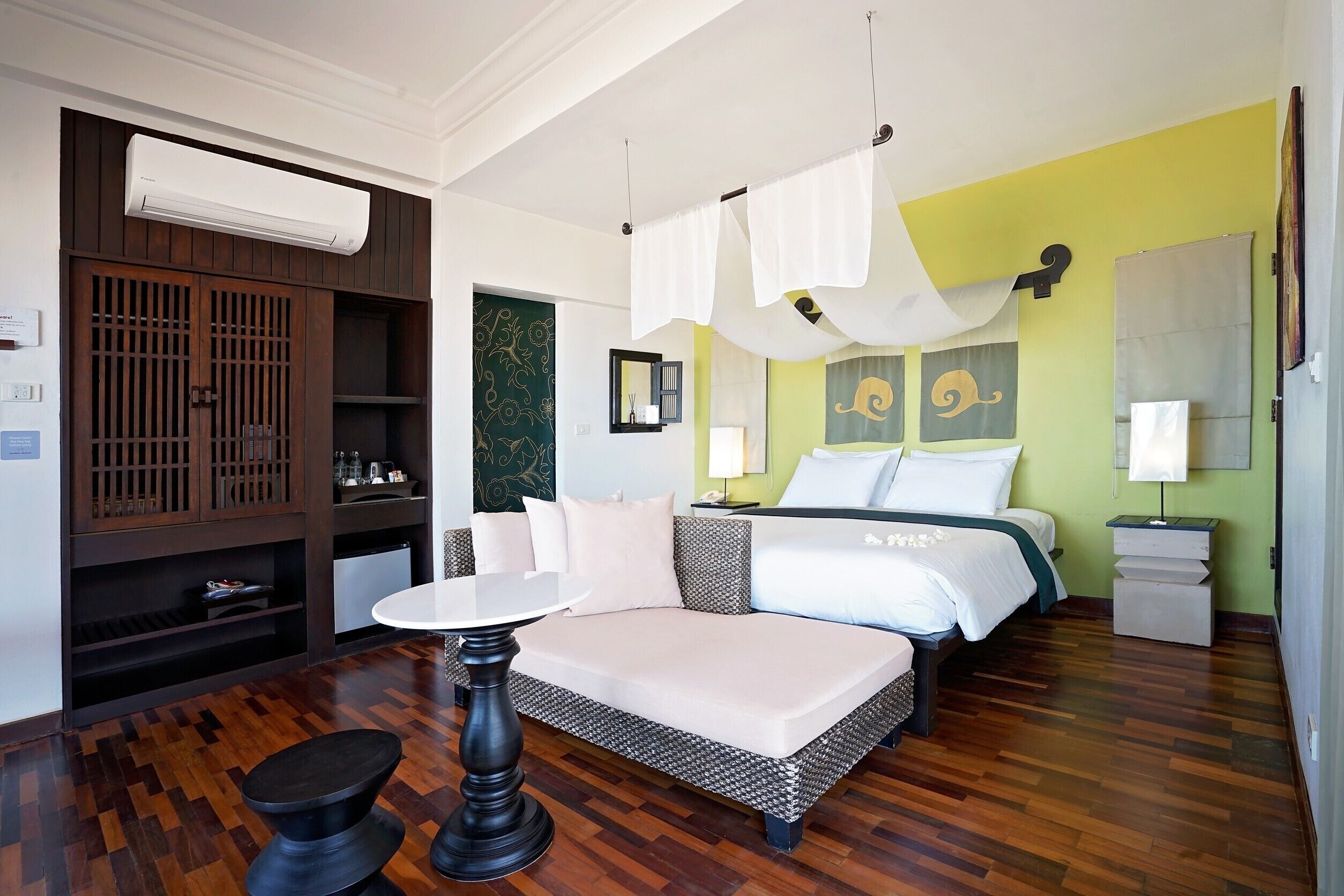 A hotel room with a bed, bedside tables, a small table, a wardrobe, and an air conditioning unit.