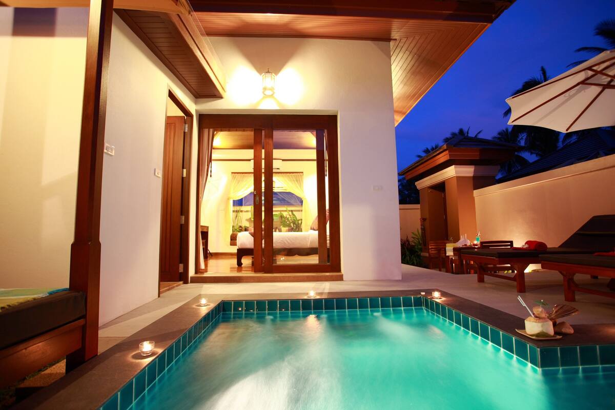 A pool area with a view of a room through a glass door, a lounge chair, and a table with a drink.