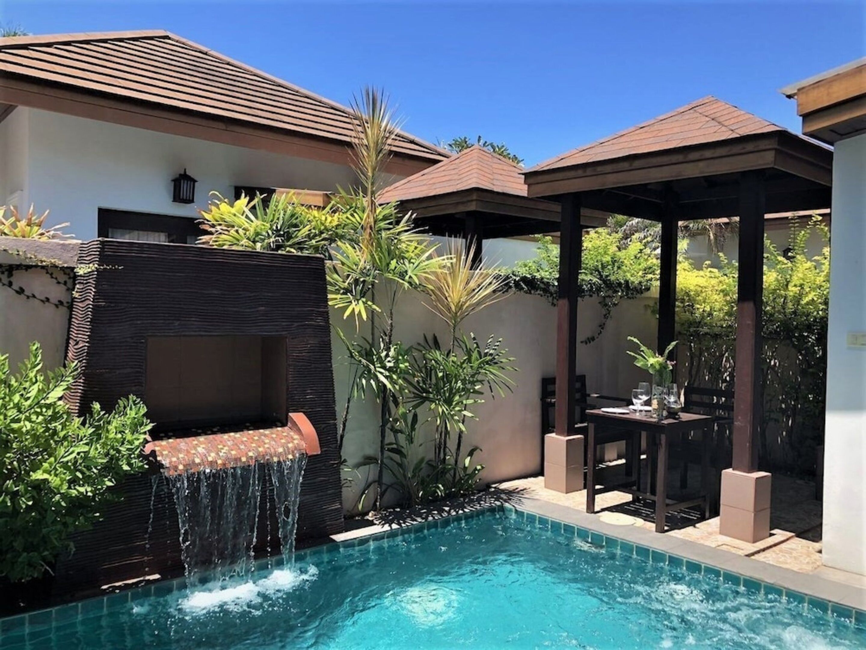 A swimming pool with a waterfall feature, surrounded by lush greenery and a covered seating area.