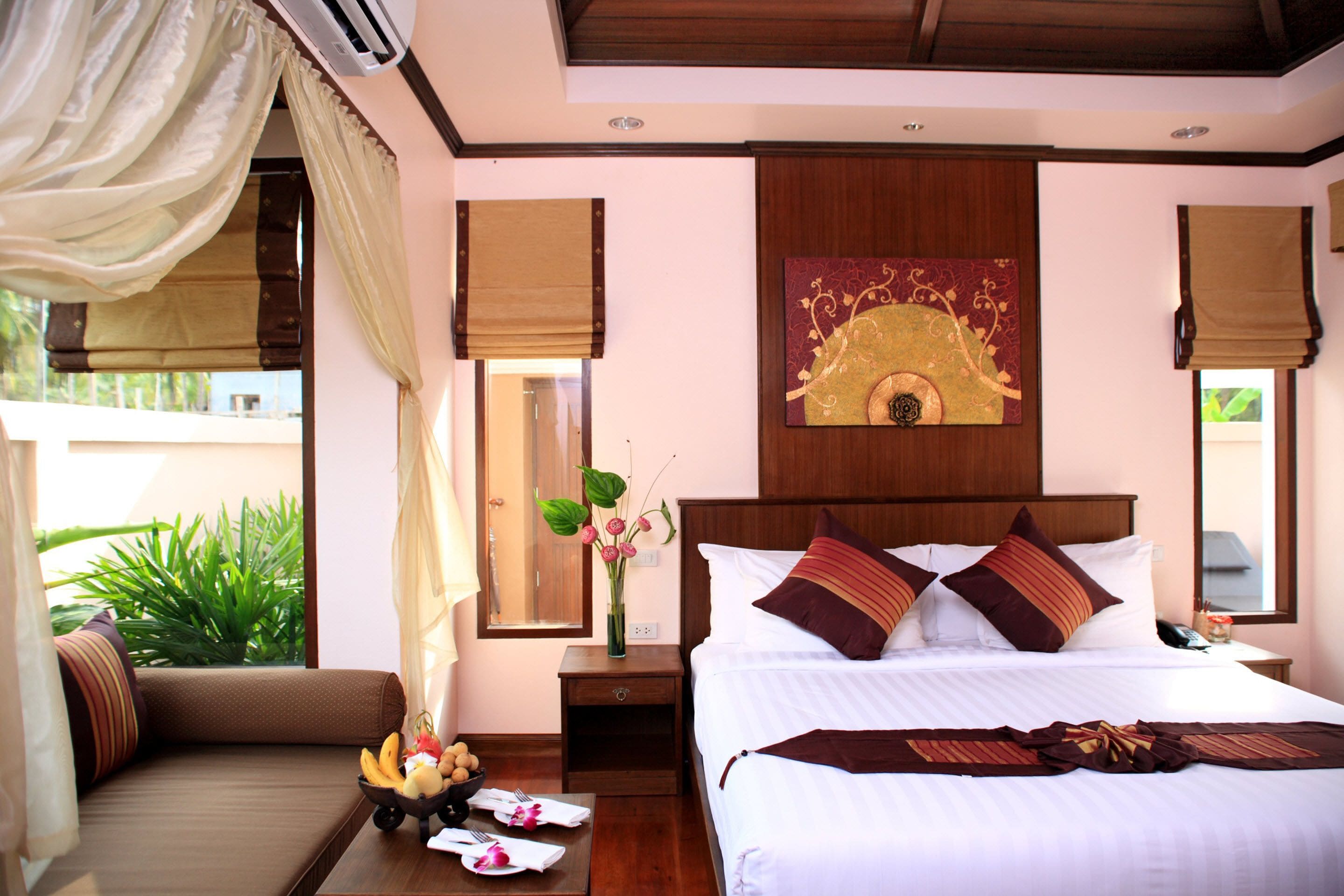 A hotel room with a large bed, a bedside table with a vase of flowers, a small table with fruits and a bowl of snacks, and a view of greenery outside.