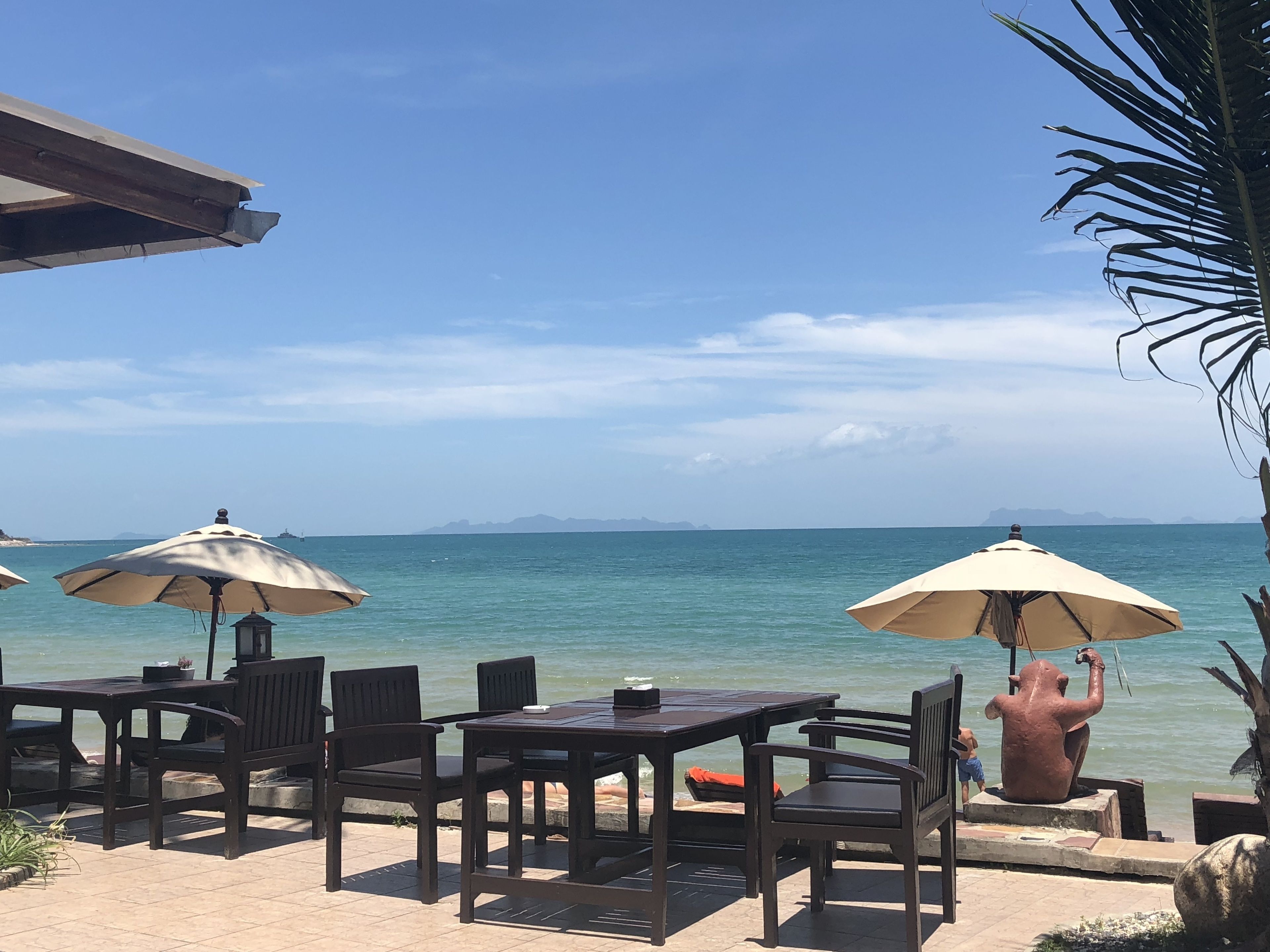 An outdoor seating area with tables and chairs, umbrellas, and a statue by the seaside.