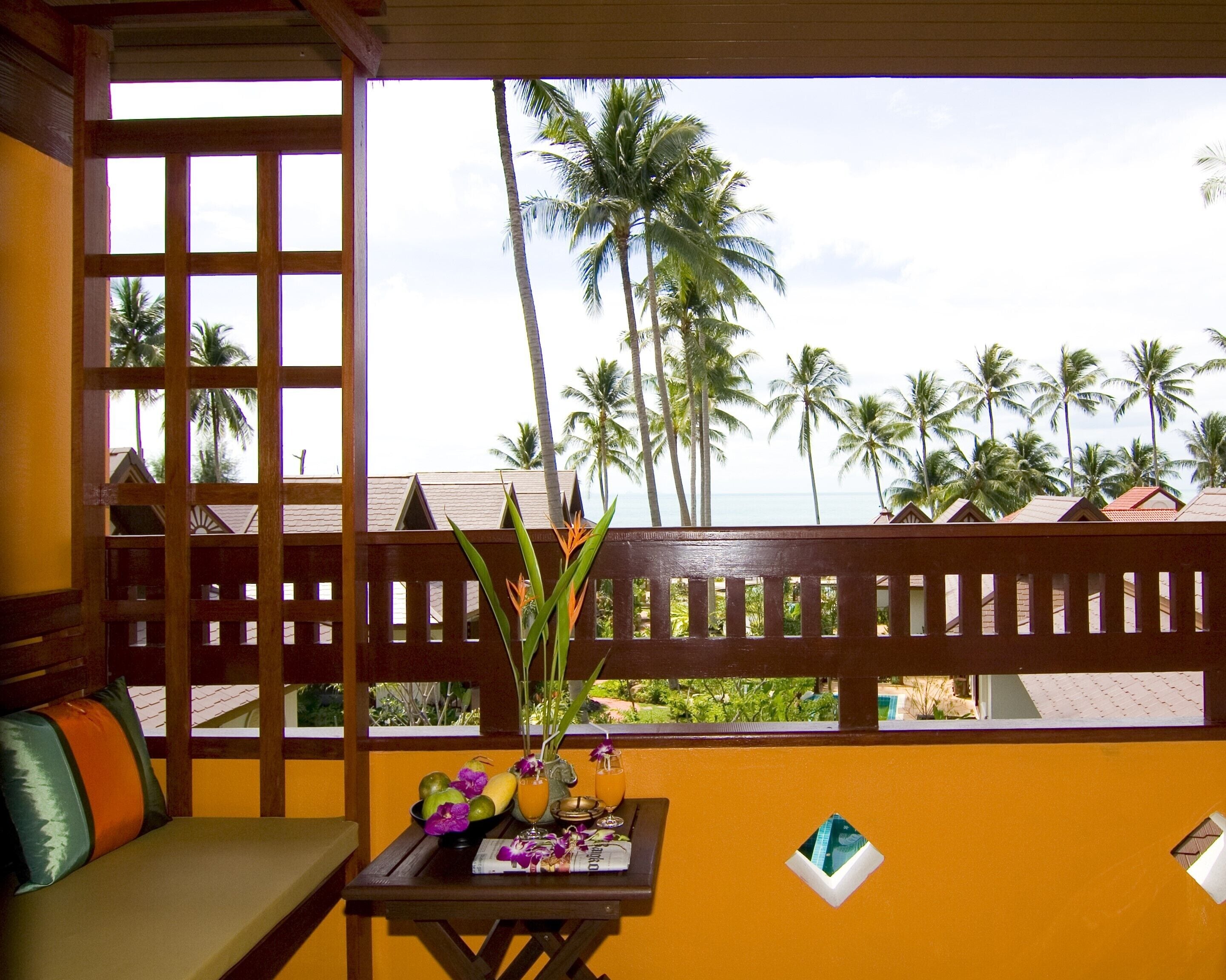 A room with a wooden bench, a small table with a newspaper, a vase with flowers, and a view of palm trees and a beach.