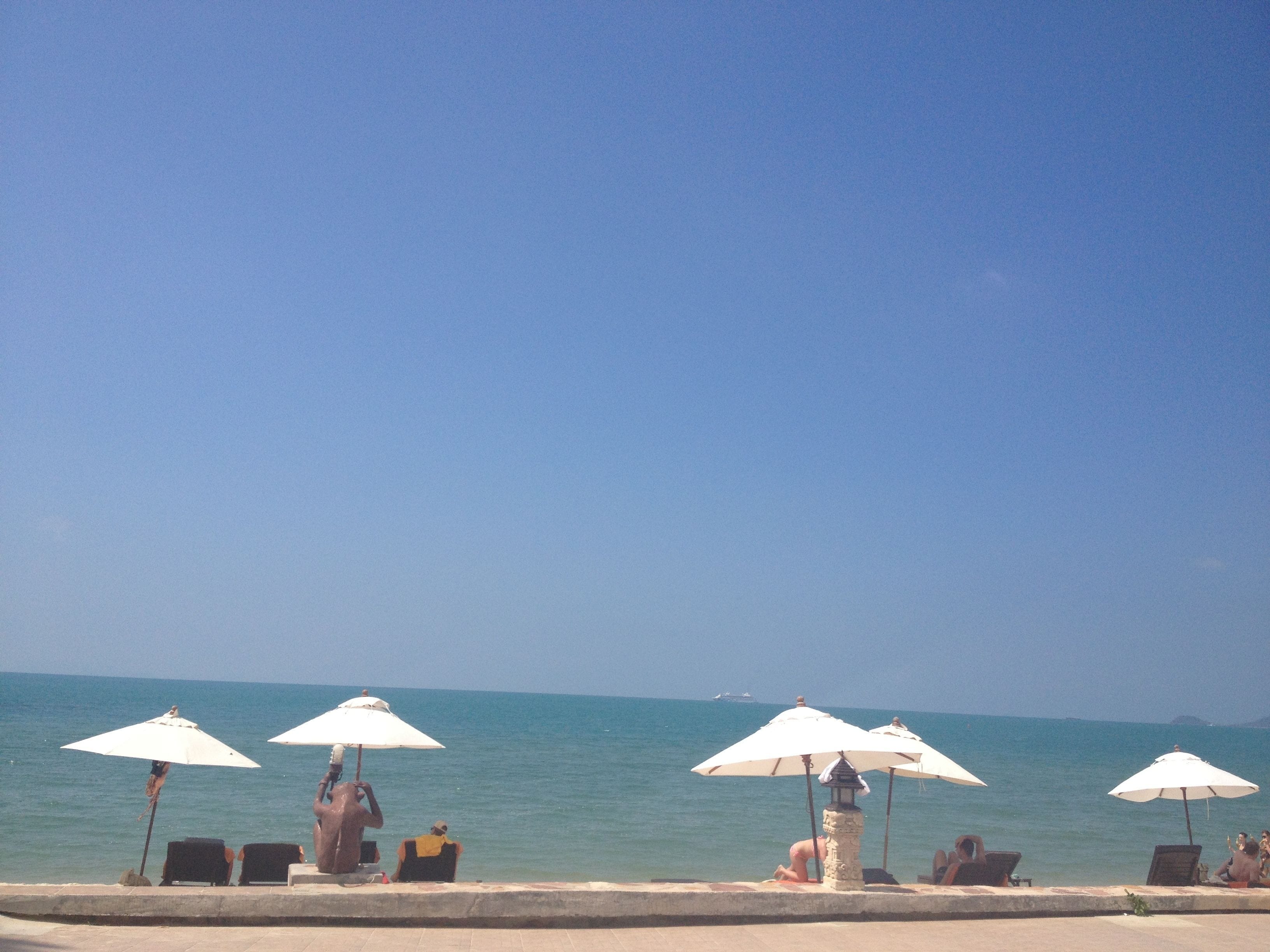 A beachside area with multiple white umbrellas providing shade, a stone wall in the foreground, and the ocean in the background.