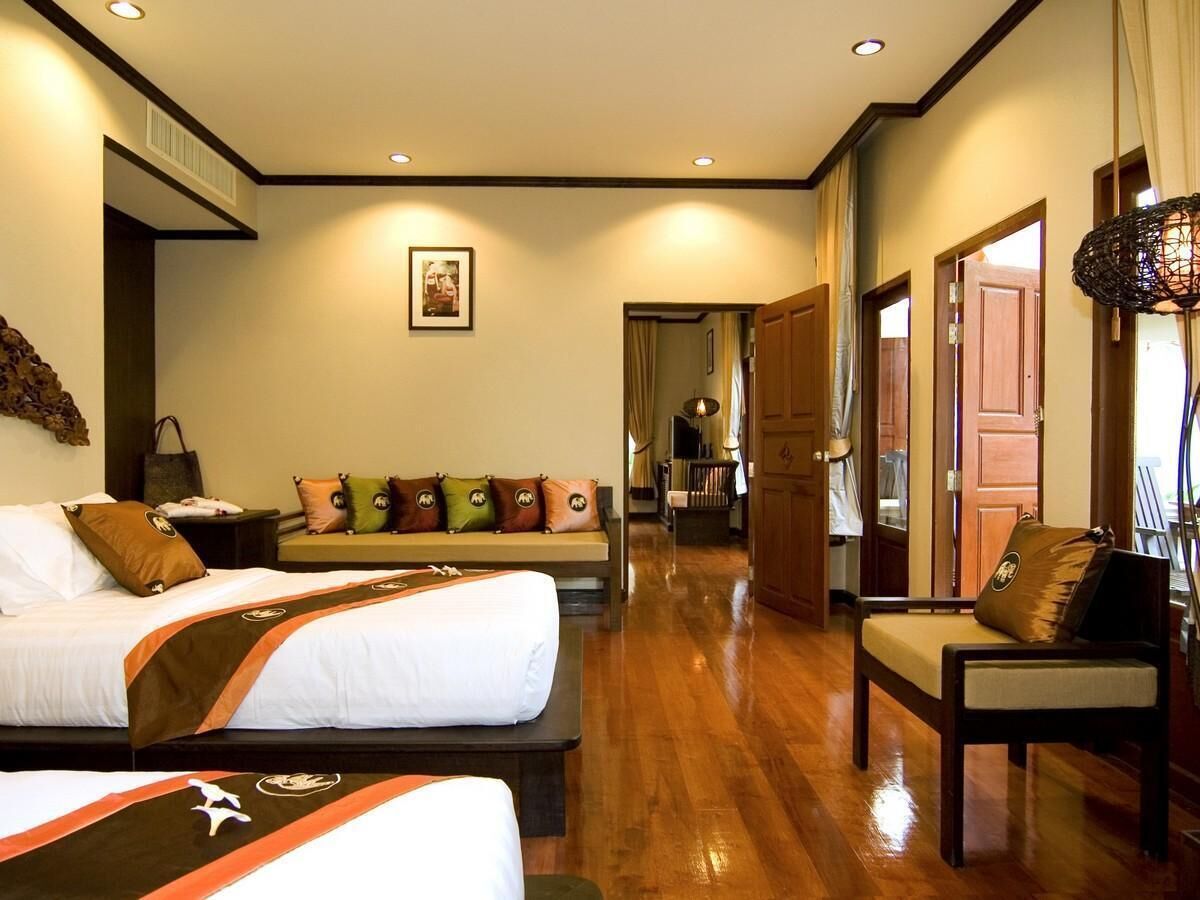 A hotel room with two beds, a sofa, a chair, wooden flooring, and a hallway leading to a bathroom.