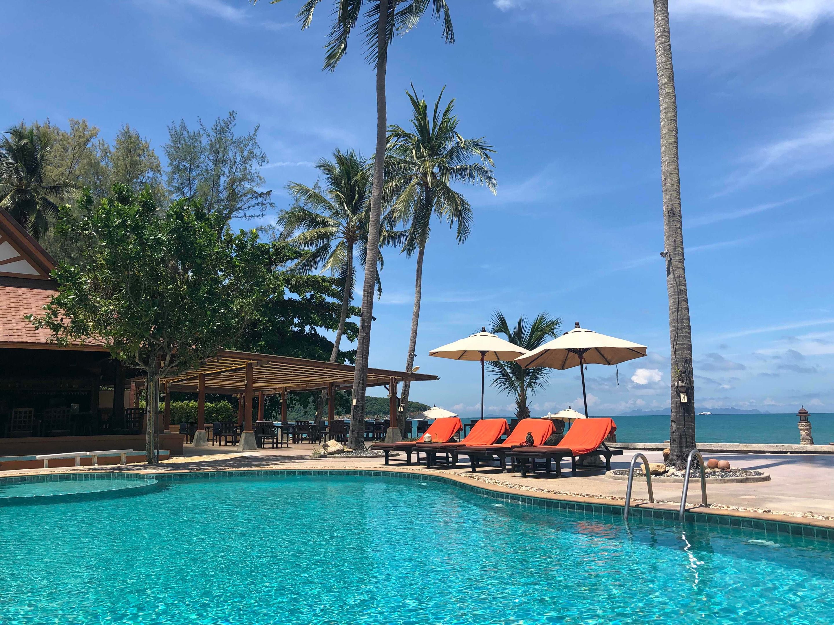 A pool area with lounge chairs and umbrellas, surrounded by palm trees and a building with a covered seating area.