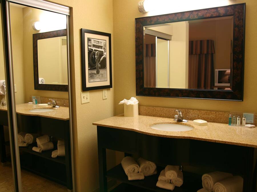 A hotel bathroom with two sinks, a large mirror, and towels neatly stacked on shelves.