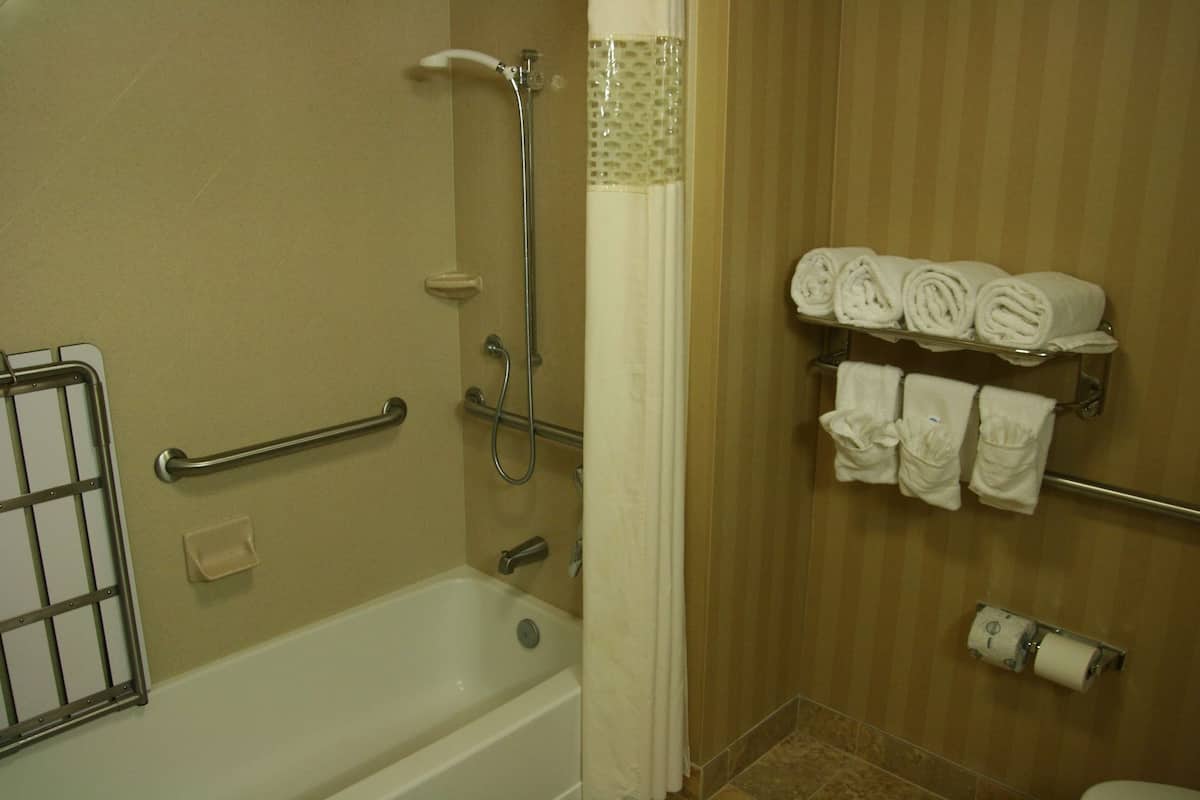 A bathroom with a bathtub, a shower, a handrail, a towel rack with towels, and a toilet paper holder.
