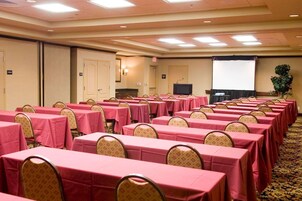 Un salón de conferencias con filas de mesas cubiertas con manteles rosas, equipado con sillas, una pantalla de proyector y un televisor.