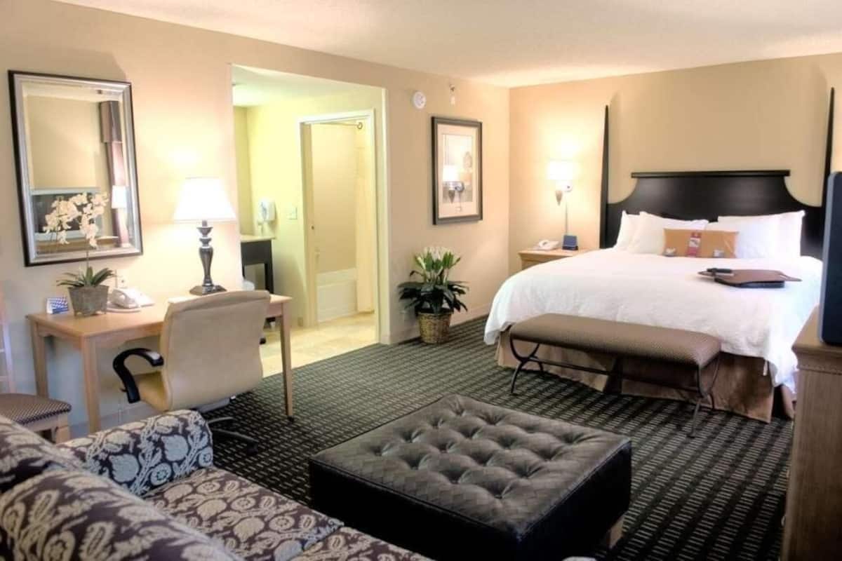 A hotel room with a bed, a desk, a chair, a sofa, a small ottoman, and a bathroom visible through an open door.