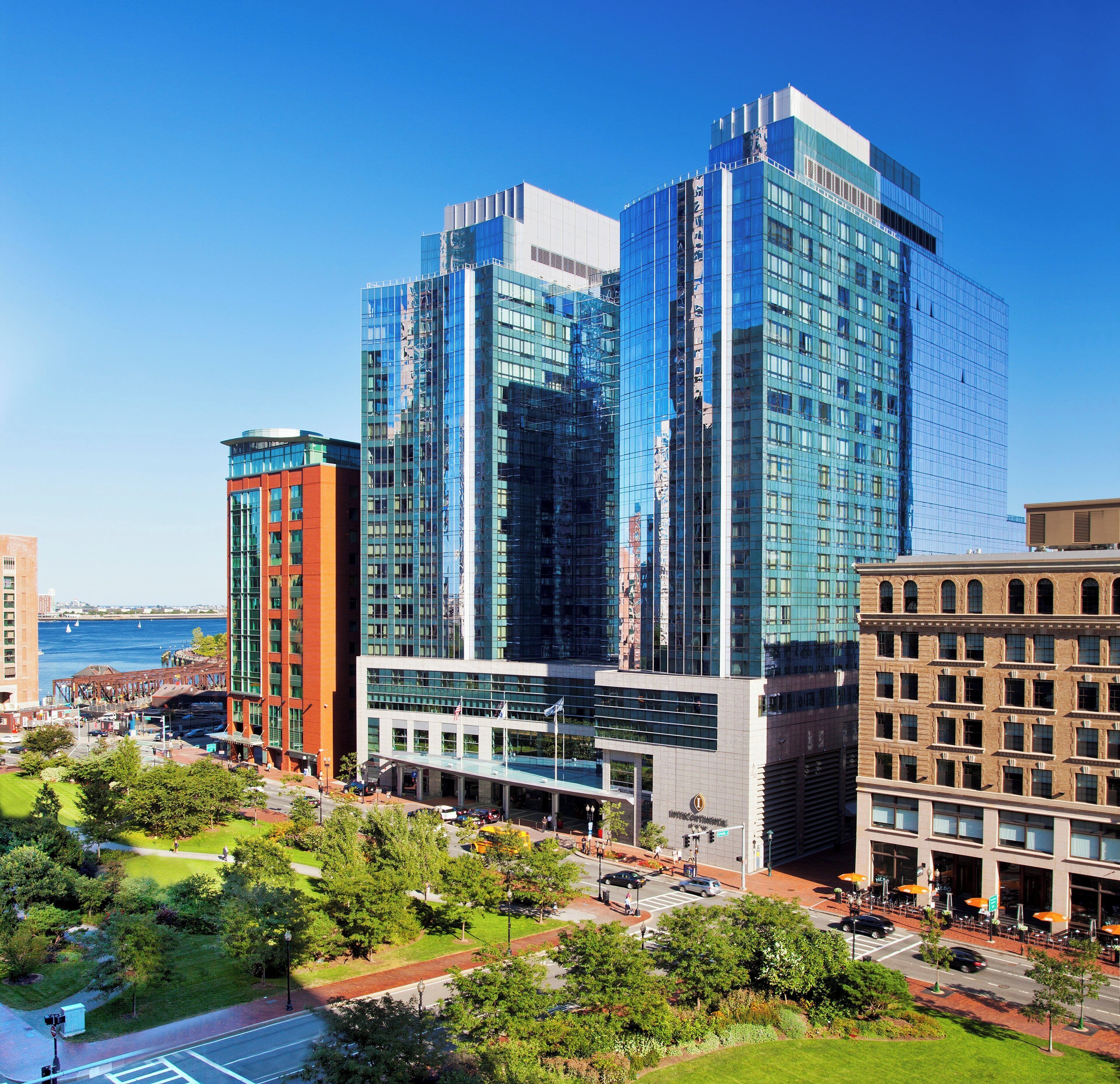 A modern building with a glass facade, surrounded by greenery and a waterfront.
