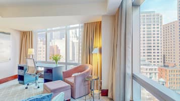 A hotel room with a desk, chair, and ottoman, large windows with curtains, and a view of city buildings.