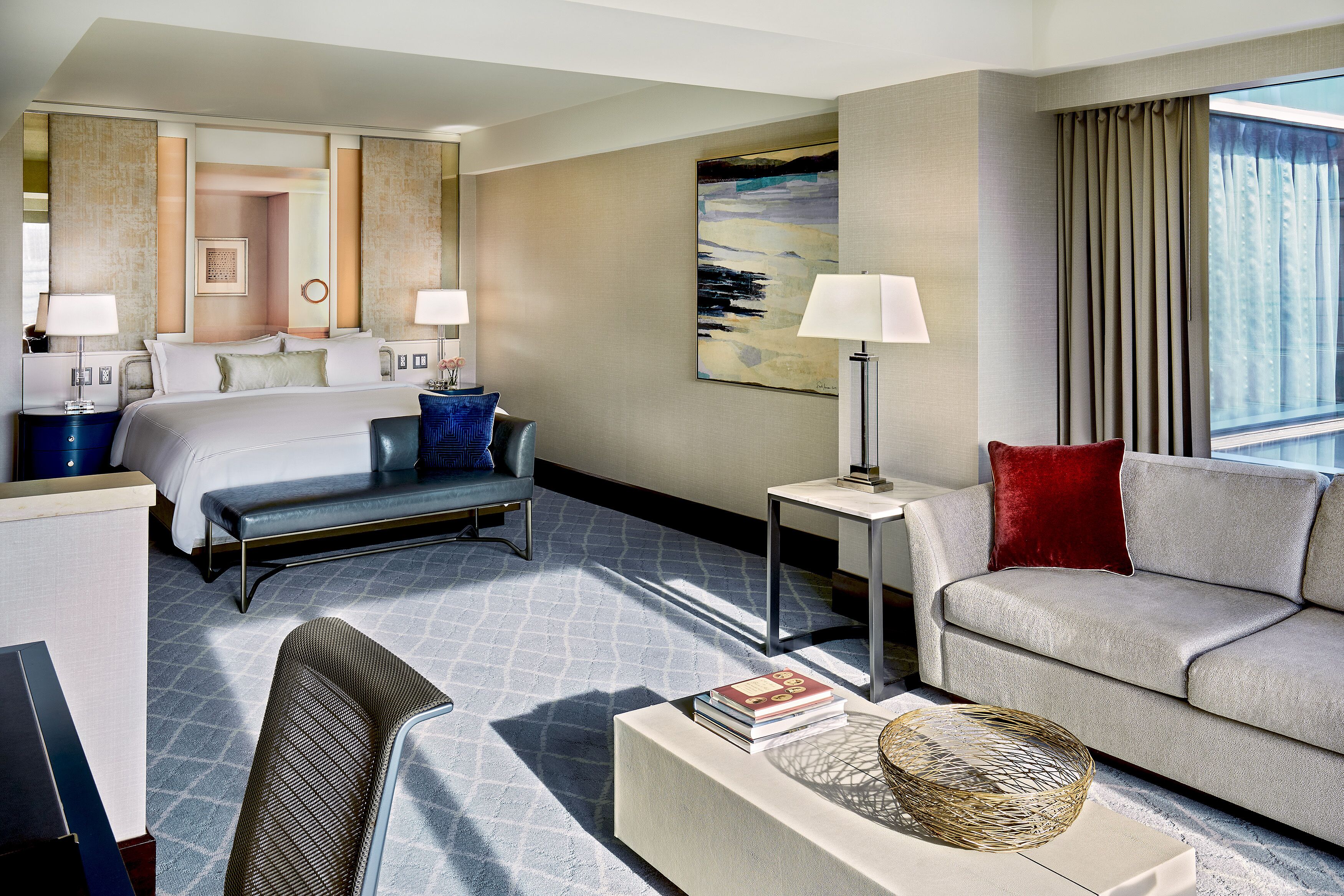 A hotel room with a bed, a sofa, a coffee table, a side table, and a large window with curtains.