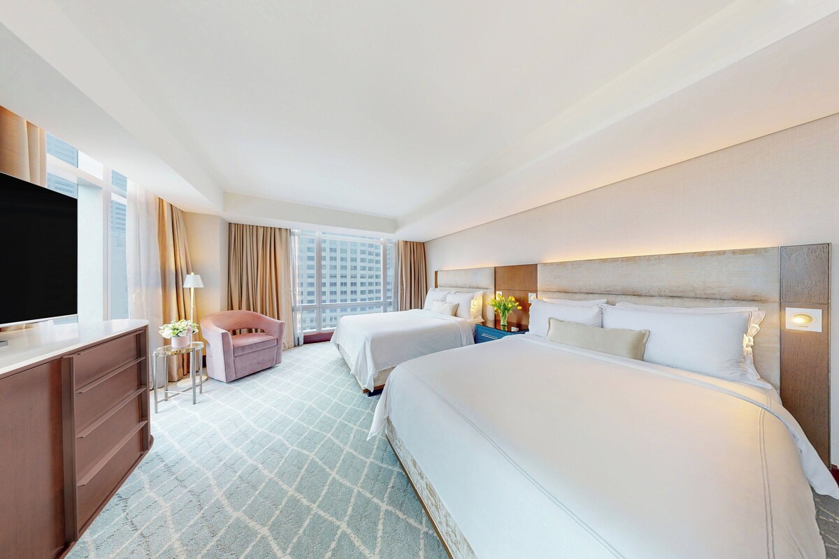 Una habitación de hotel con dos camas, un escritorio, una silla, un televisor y un ventanal grande.