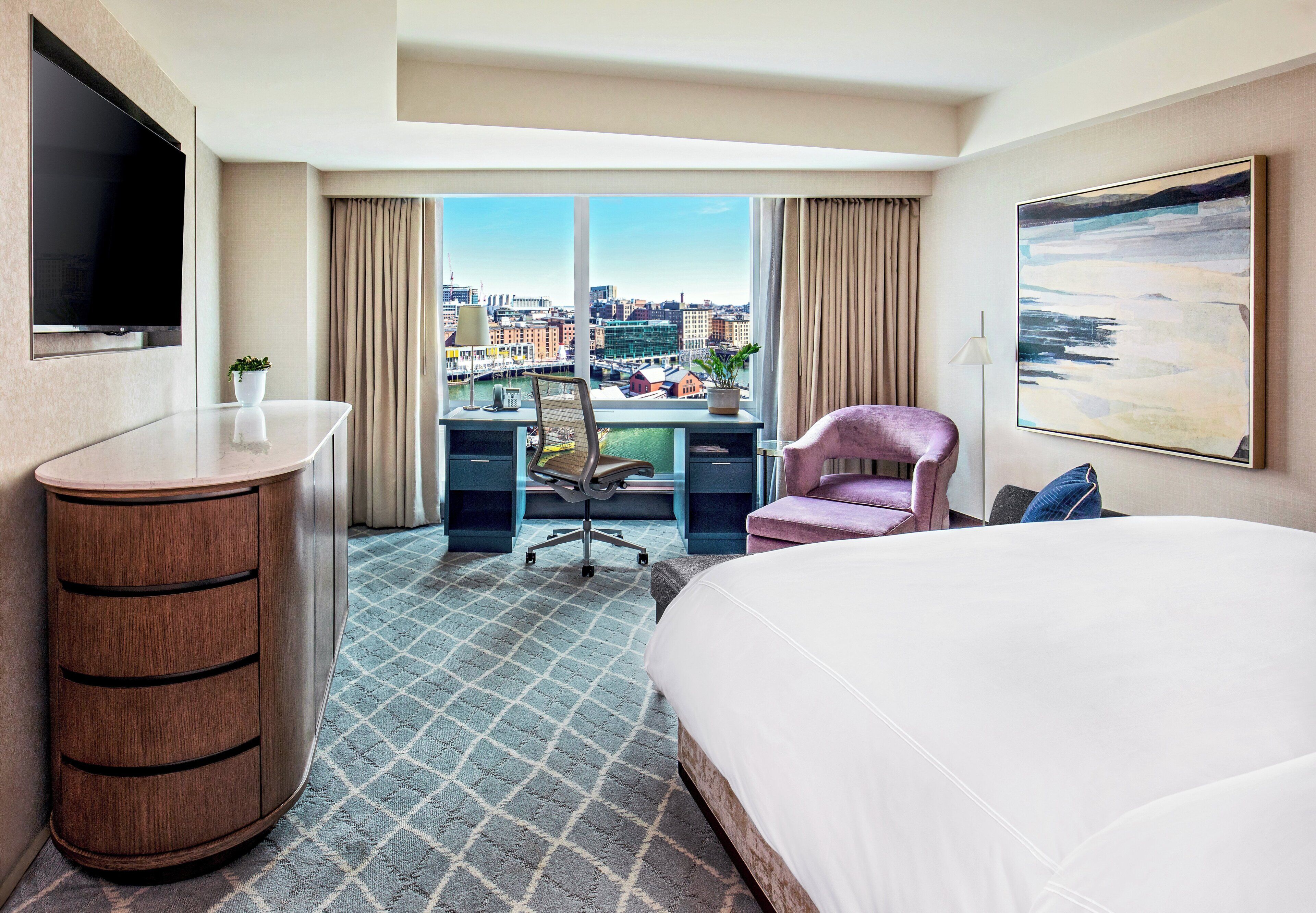 A hotel room with a bed, desk, chair, TV, and a large window with a city view.