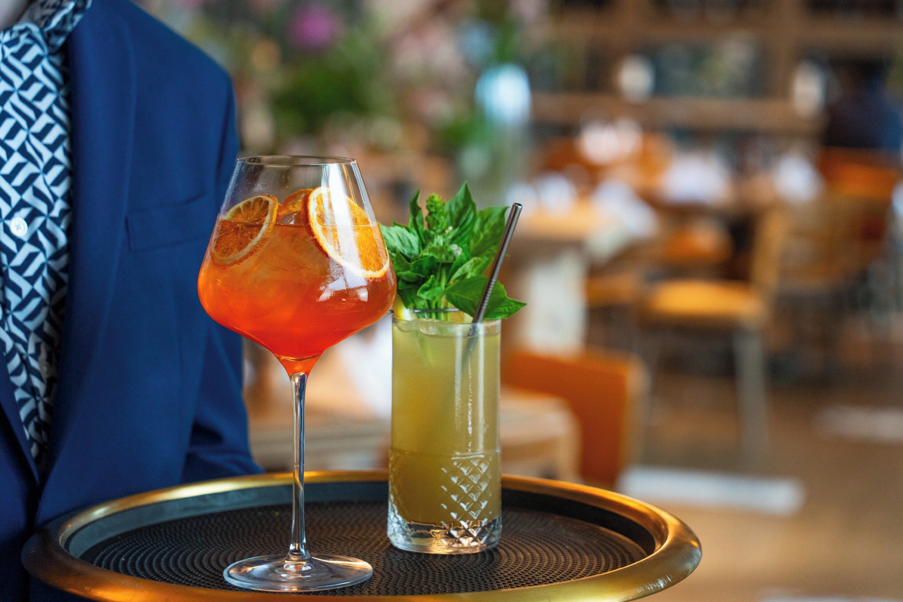 A table with two drinks, one in a martini glass and one in a highball glass, with a blurred background of a restaurant setting.