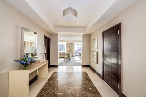 A hotel hallway with a marble floor, a reception desk with a vase of flowers, a large mirror, and a door leading to a living area with a view of the city.