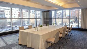 Un salón de conferencias con una larga mesa, sillas y grandes ventanales que ofrecen vistas a la ciudad.