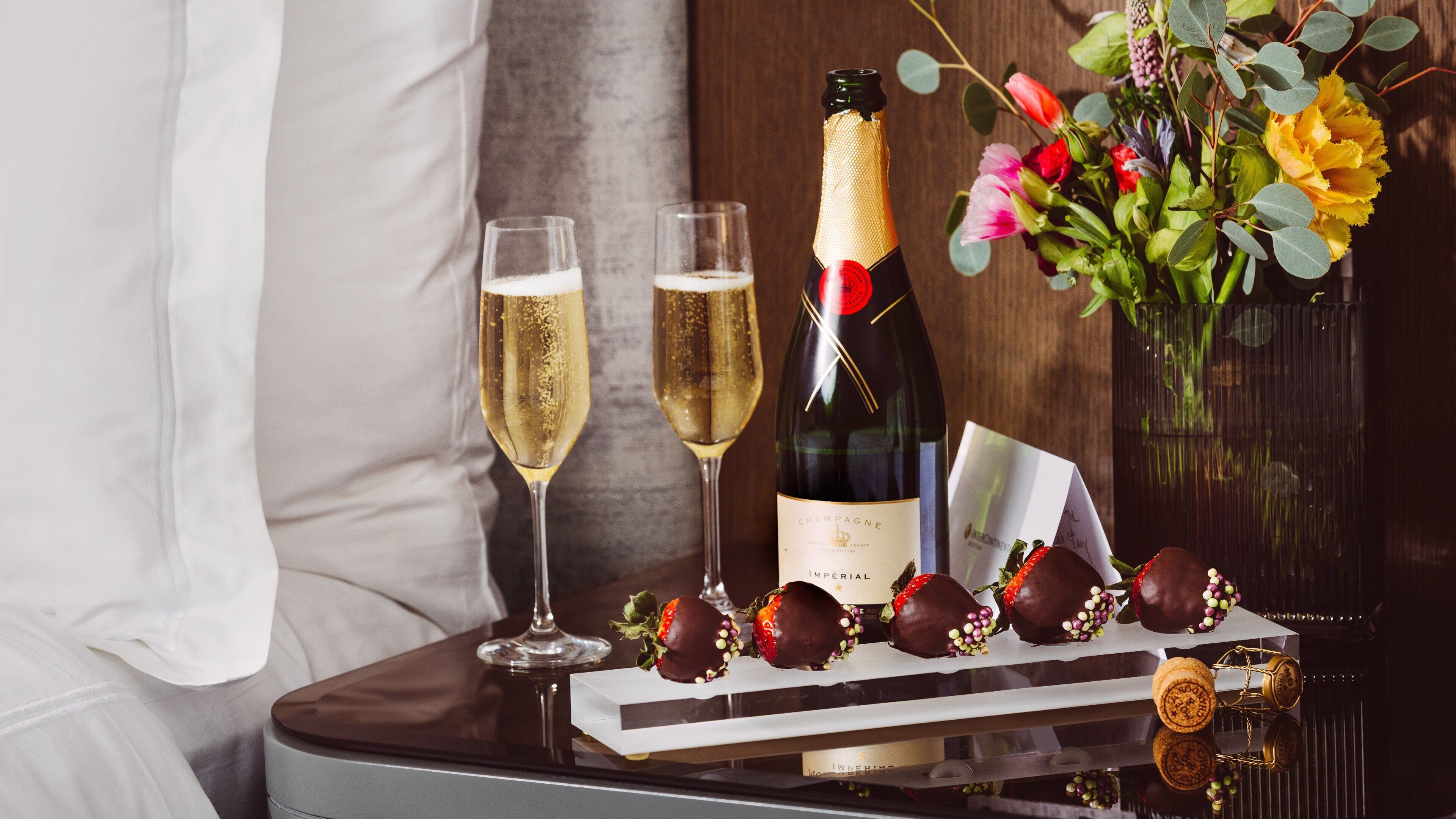 A bottle of champagne, two glasses, chocolate-covered strawberries, and a decorative vase on a table.