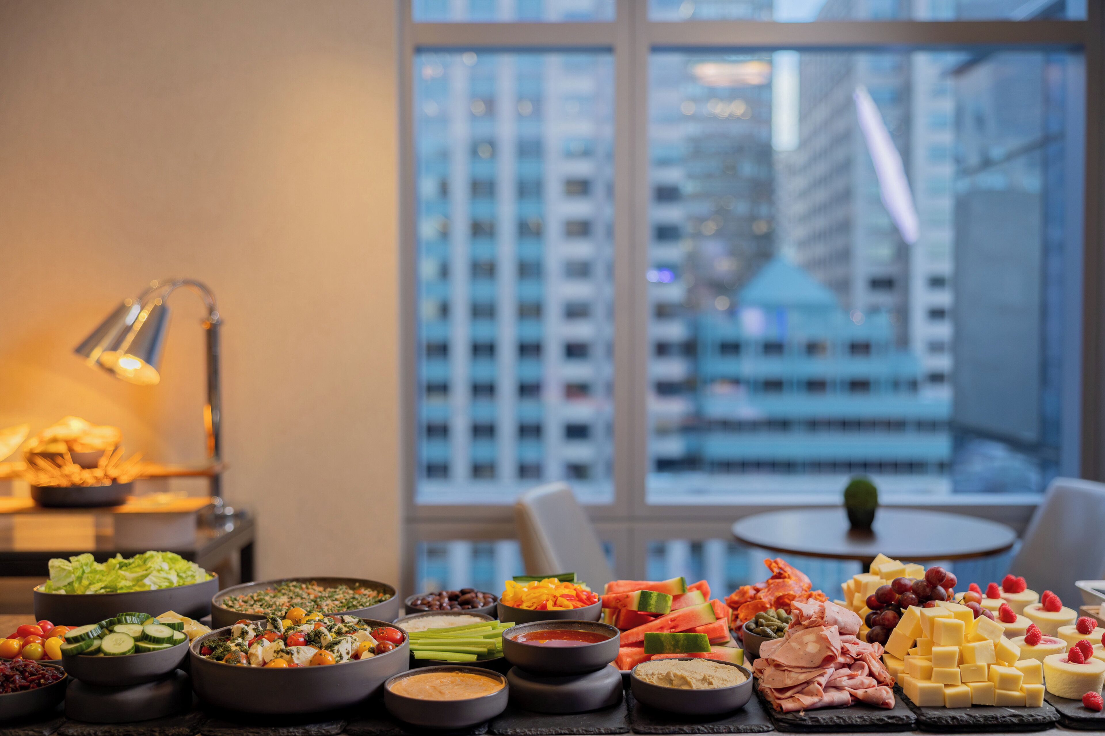 A buffet table with various food items and a city view through large windows.