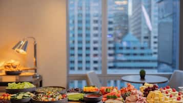 A buffet table with various food items and a city view through large windows.