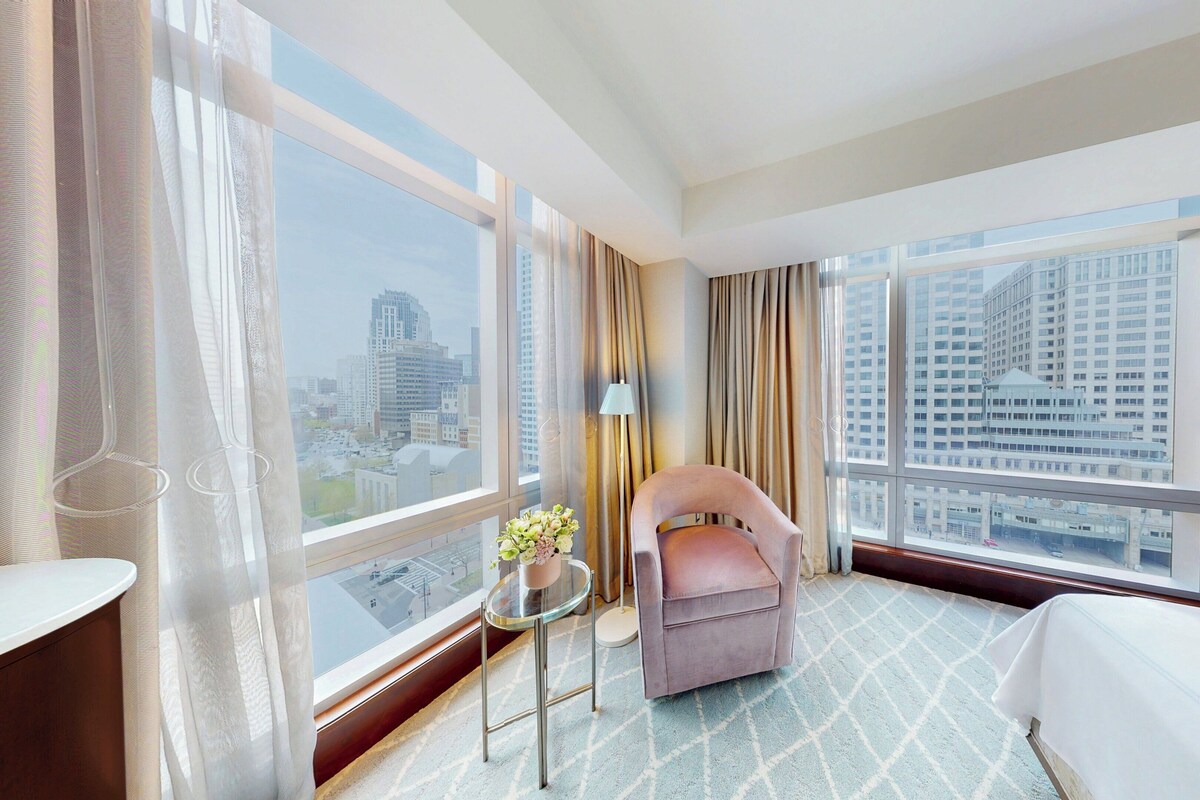 Habitación de hotel con cama, una silla, una mesita con un florero y ventanas amplias que ofrecen vista a la ciudad.