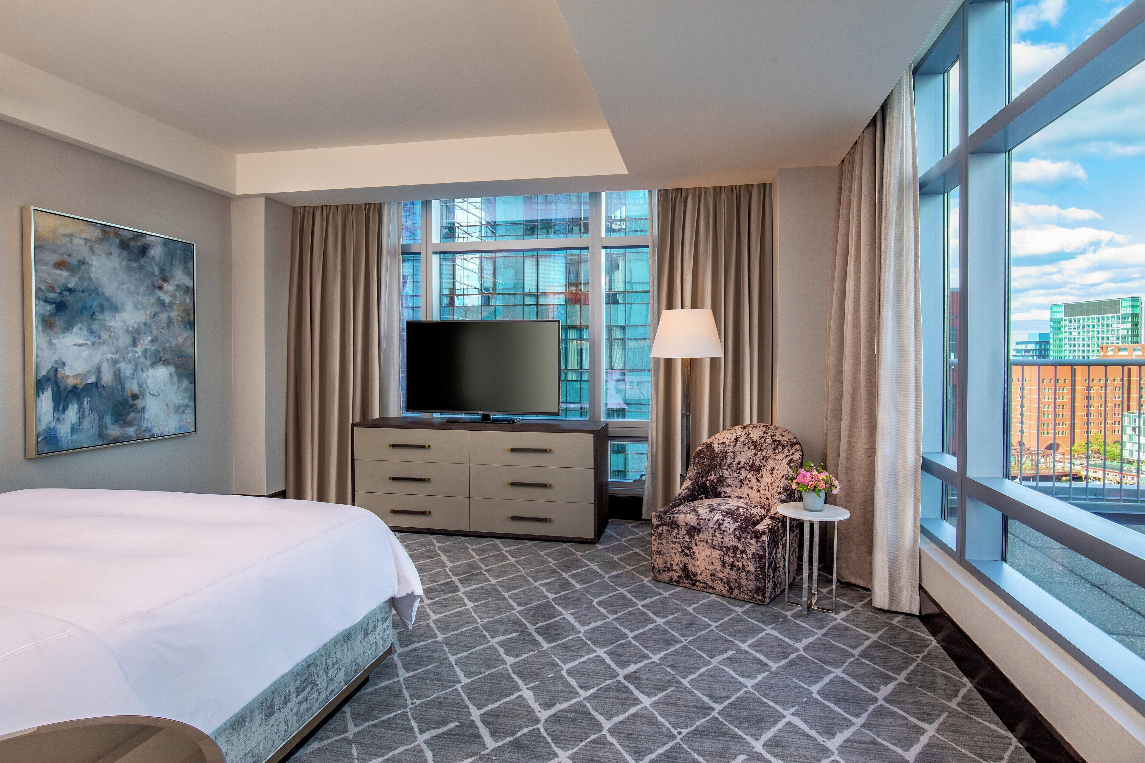 Habitación de hotel con una cama grande, un televisor de pantalla plana, un sofá, una mesita con un florero y vistas a la ciudad a través de amplias ventanas.