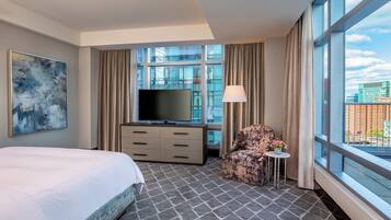 Habitación de hotel con una cama grande, un televisor de pantalla plana, un sofá, una mesita con un florero y vistas a la ciudad a través de amplias ventanas.