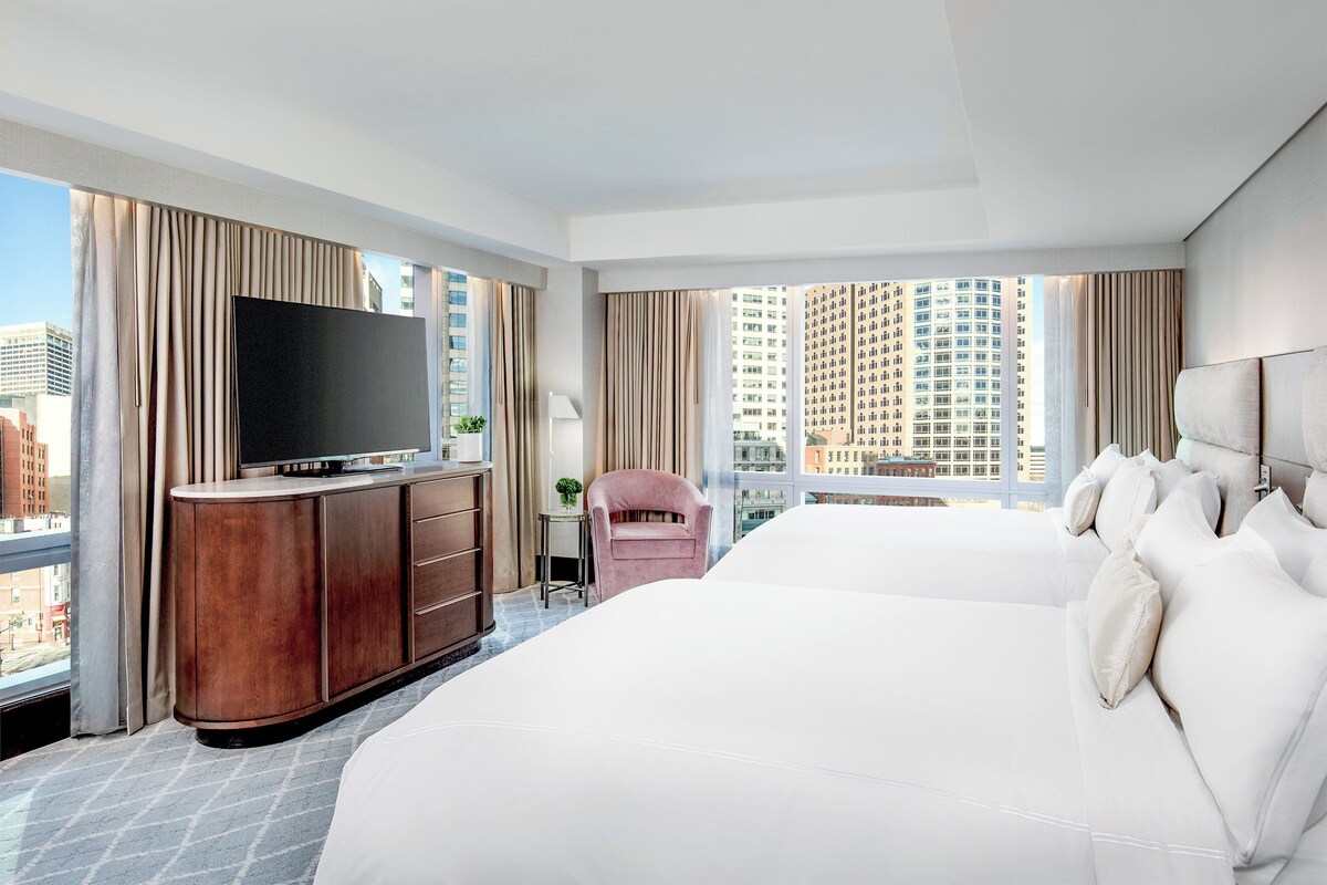 Habitación de hotel con una cama grande, un televisor sobre un mueble de madera, una silla rosa y vistas a la ciudad.
