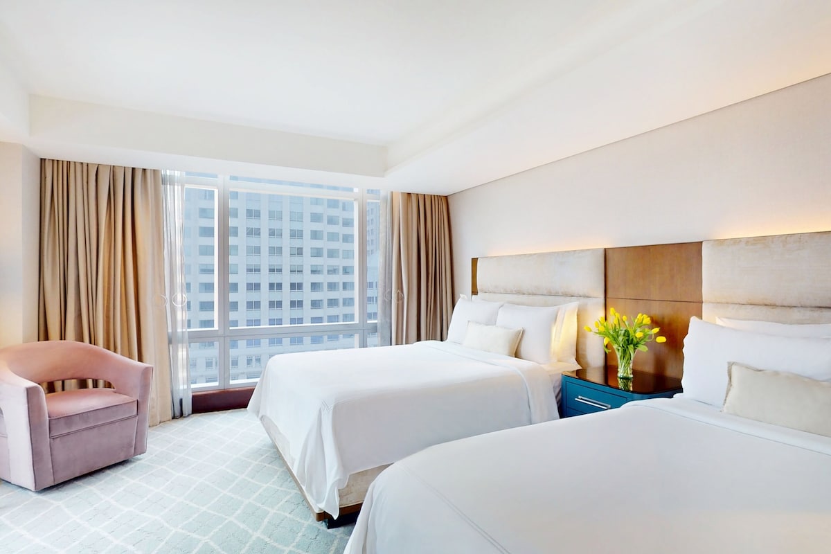 A hotel room with two beds, a chair, a nightstand with a vase of flowers, and a large window with curtains.