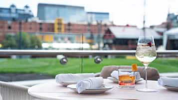 Tavolo apparecchiato per due, con un bicchiere di vino bianco e un piccolo bicchierino di liquido ambrato, probabilmente un cocktail, su una terrazza con vista sulla città.