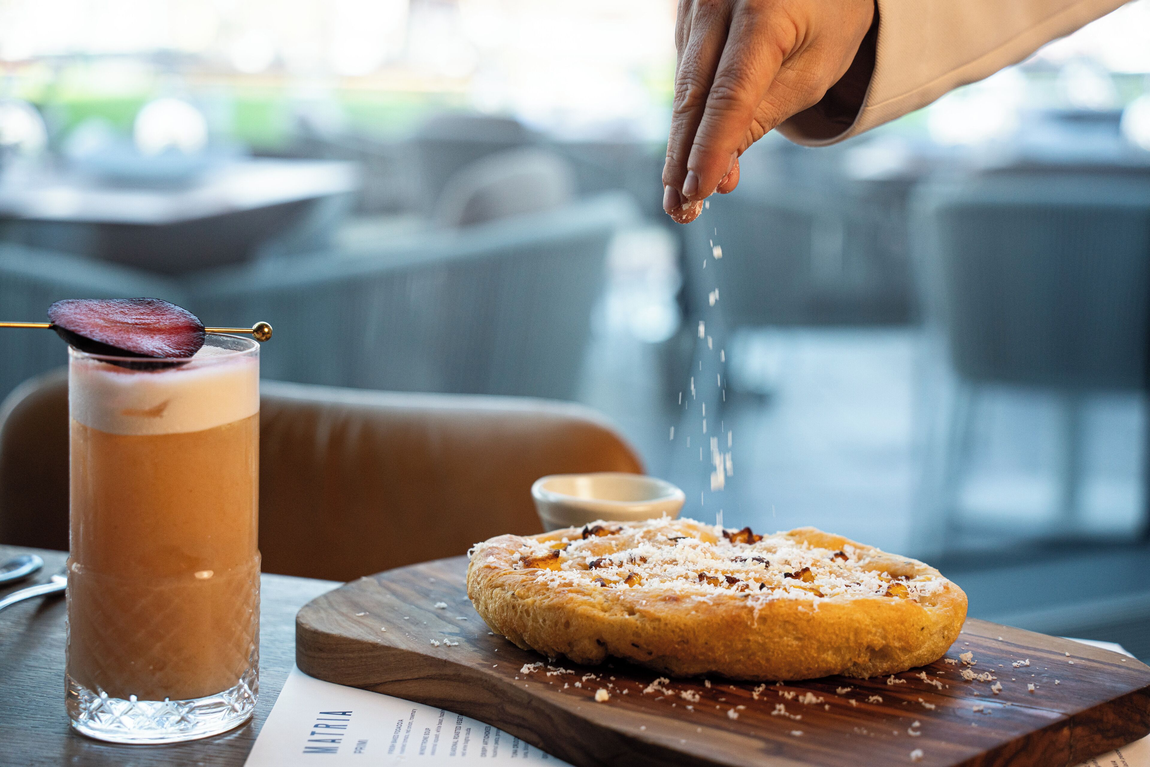A hand sprinkling salt over a dessert on a wooden board, next to a glass of a layered beverage.