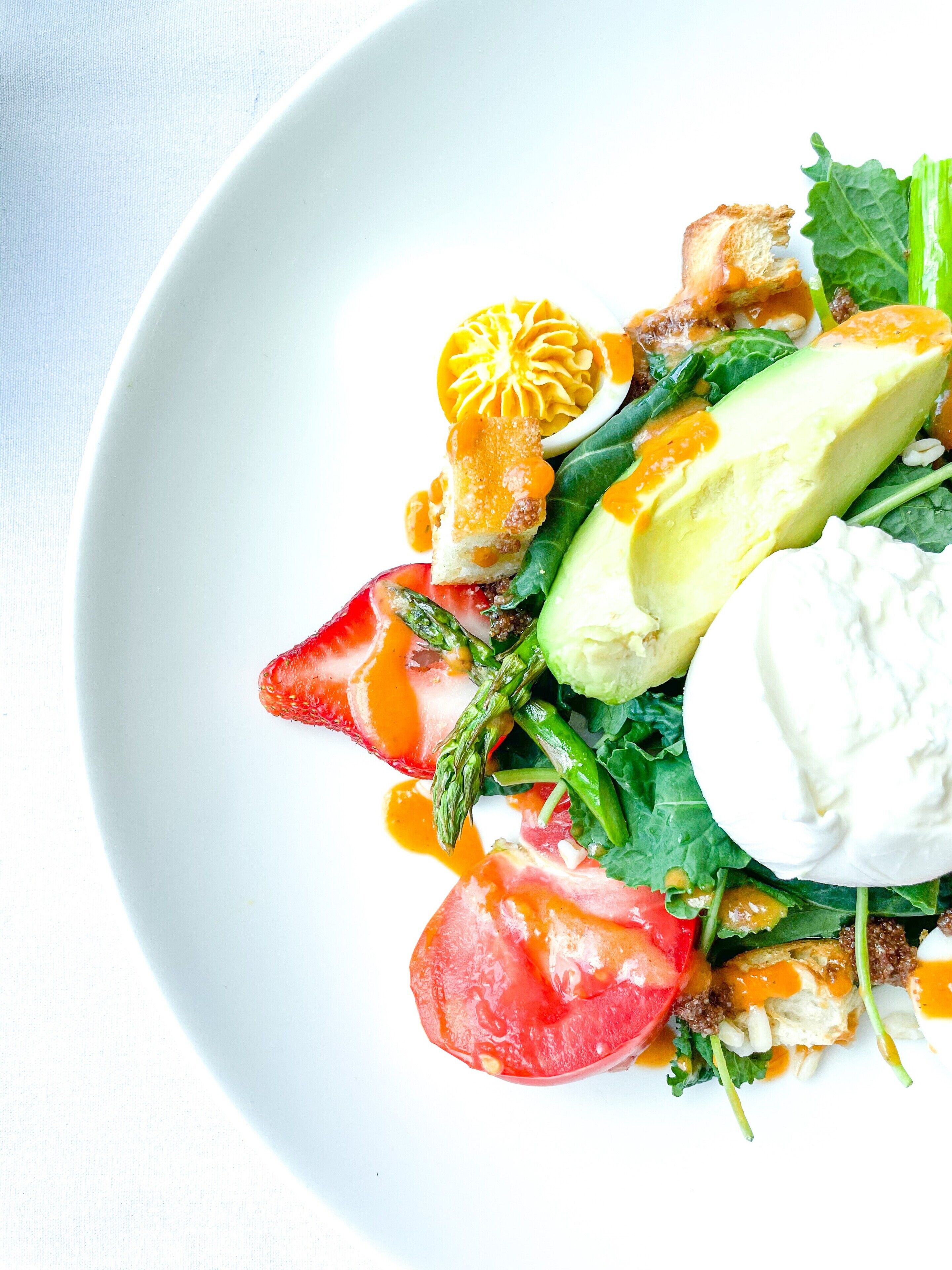 A white plate with a salad consisting of avocado, tomato, asparagus, and a poached egg.
