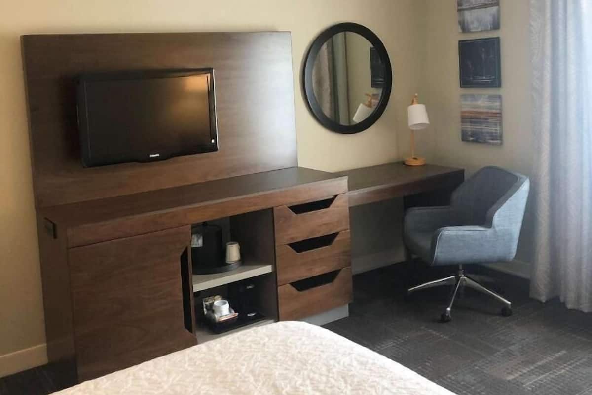 A hotel room with a bed, a desk with a flat-screen TV, a chair, and a round mirror.