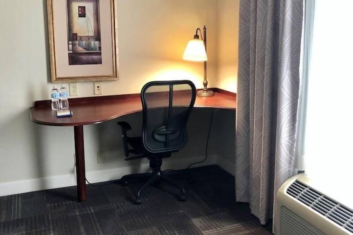 A hotel room with a desk, chair, lamp, framed picture, and a window with curtains.