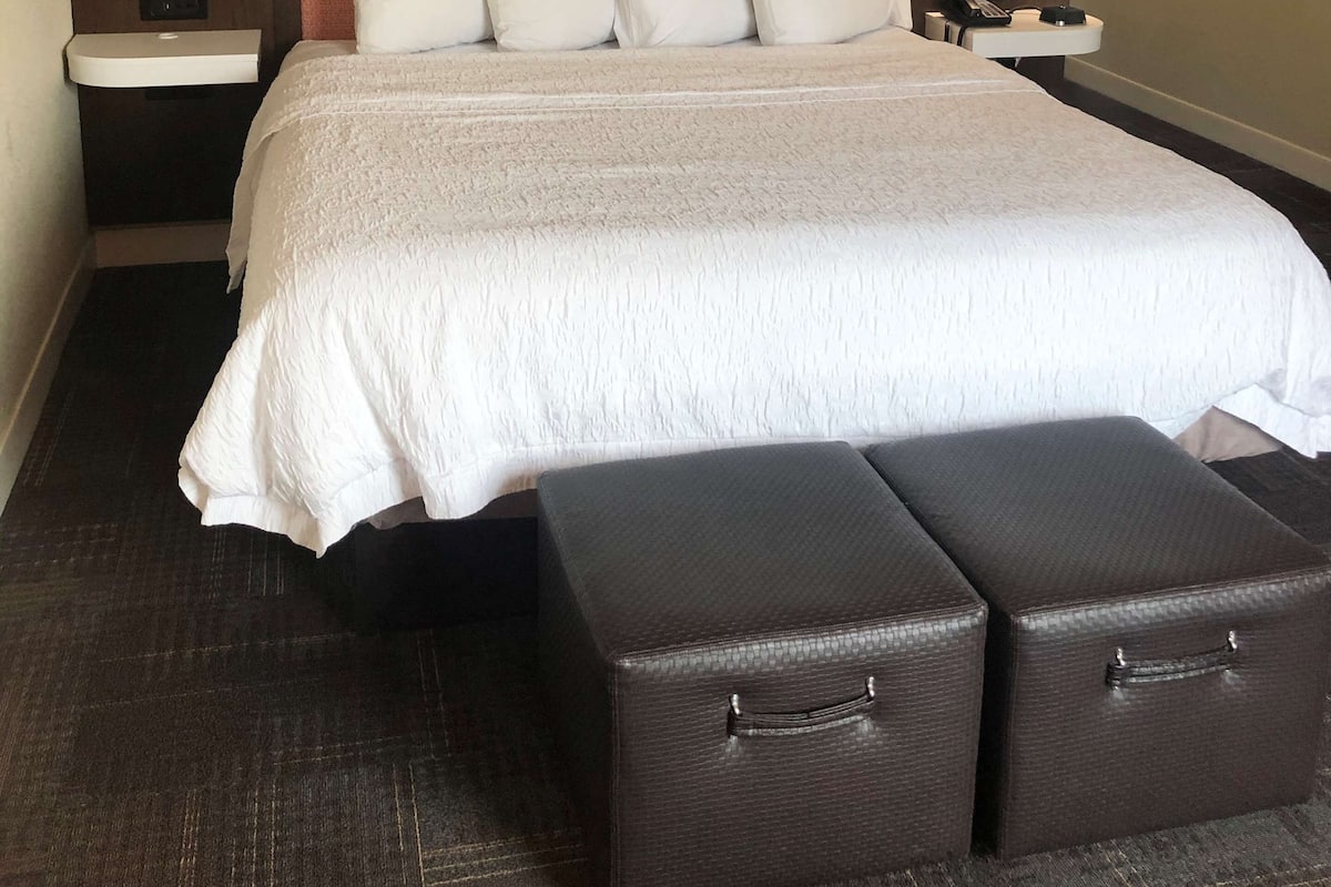 A hotel room with a large bed, a nightstand, a lamp, and two storage bins.