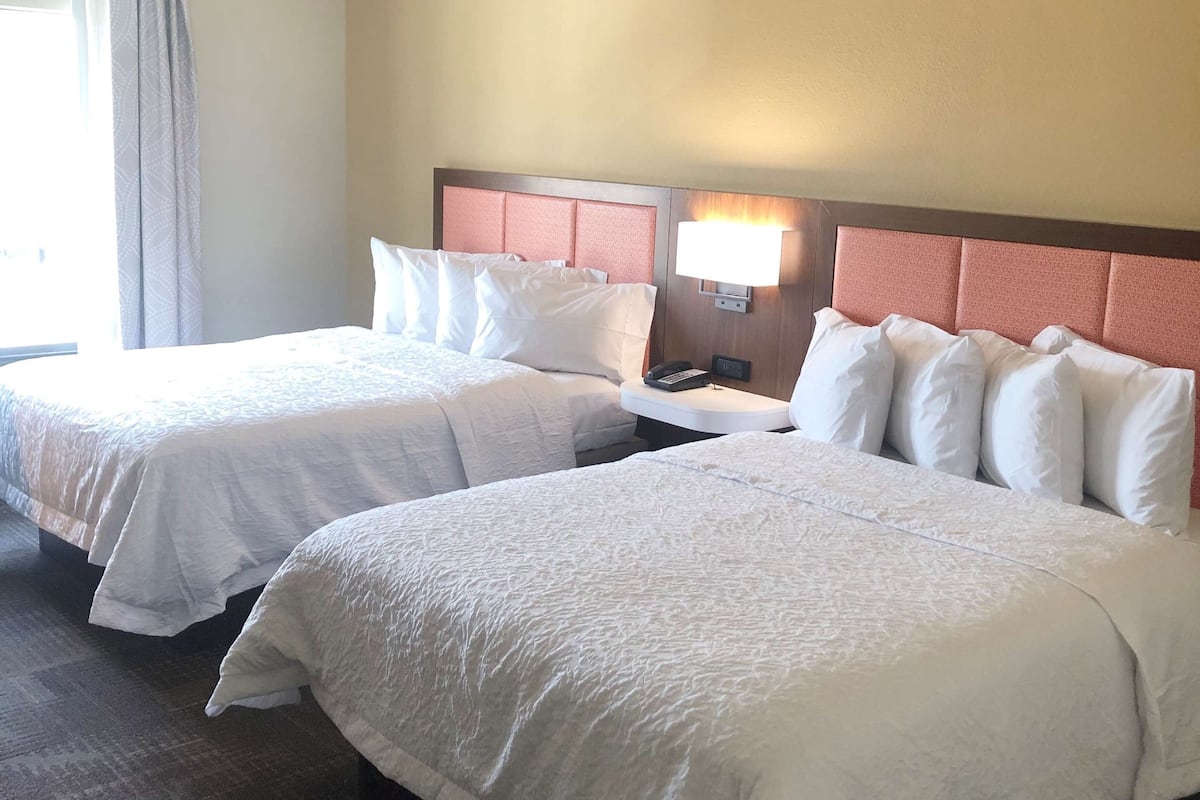 A hotel room with two beds, a nightstand with a lamp, and a window with curtains.