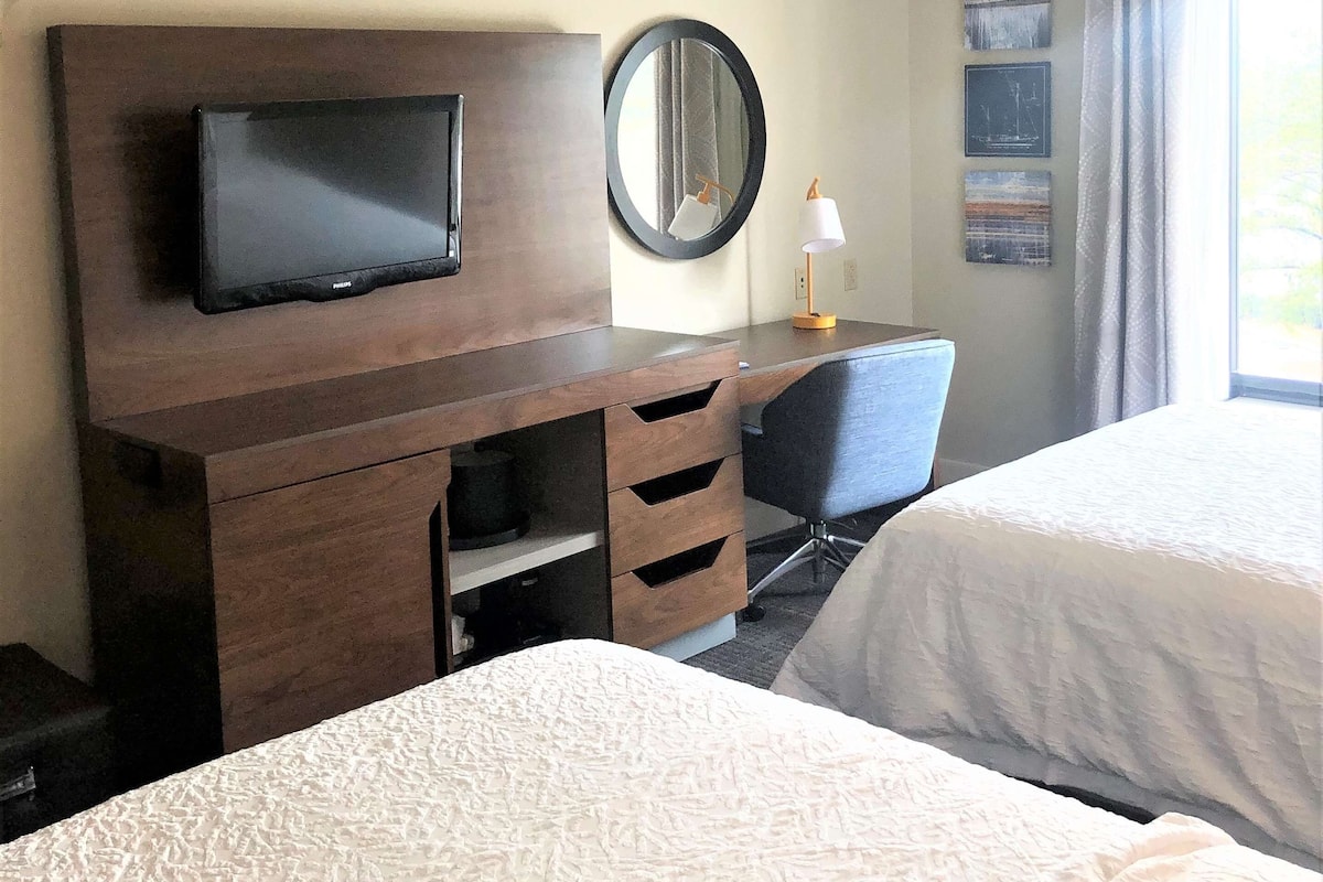 A hotel room with two beds, a desk with a television, a mirror, and artwork on the wall.