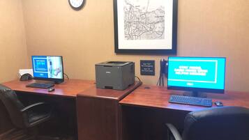 Un salón de conferencias con dos escritorios, cada uno equipado con una computadora, teclado y ratón. Una impresora se encuentra sobre el escritorio central. Un reloj y un mapa enmarcado están colgados en la pared.