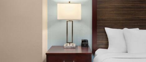 A neatly made bed with white linens, a bedside table with a lamp, and a small electronic device on the table.