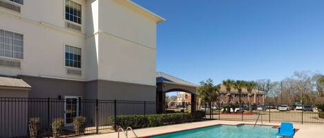 A pool area with a clear blue pool, a blue lounge chair, and a building with multiple windows and a covered walkway.